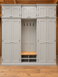 A large light gray wooden entryway cabinet and upper cabinet, a central coat rack with hooks, a wooden bench seat, and open storage shelves below, shown against a wood-paneled wall and wood floor.