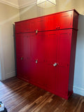 A large red six-door storage cupboard with a ironing board space, positioned on a room with wooden flooring.