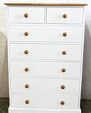 White 2 over 5 chest of drawers with wooden drawers and knobs against a white brick wall.