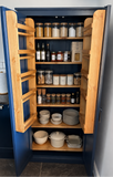 A tall navy blue pantry cabinet with both doors swung open, showing wooden shelves stocked with glass canisters, bottles, jars, plates, and cookware in a kitchen setting.