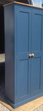 A blue hallway storage cupboard with coat hooks and three shelves, made from pine wood, with its two doors closed.